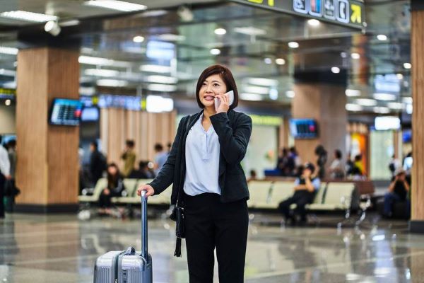 Mujer de negocios habla por teléfono celular mientras arrastra su maleta de cabina con ruedas por el suelo de un aeropuerto.