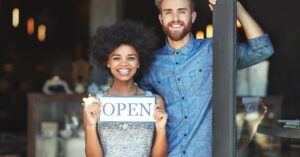 Smiling shop owners holding Open sign at doorway.